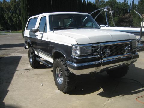 1989 Ford Bronco - Three-quarter front view of a lifted white-over-dark-blue Ford Bronco with hardtop, freshly washed on a concrete pad with a wakeboard-tower…