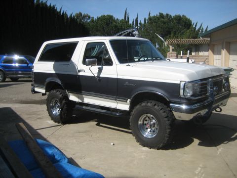 1989 Ford Bronco - Passenger-side front three-quarter view of a lifted white-over-gray Ford Bronco with hardtop, Bronco fender script, chrome grille guard and…