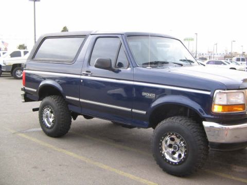 1995 Ford Bronco - This blue 1995 Ford Bronco is shown from a 3/4 front angle, with another white SUV visible in the background.