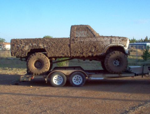 1995 Ford Bronco - A heavily mud-caked pickup truck is transported on a dual-axle trailer under a clear blue sky.