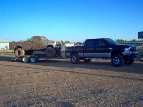 1995 Ford Bronco - A black pickup truck tows a trailer carrying a heavily mud-covered truck under a clear blue sky.
