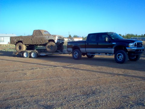 1995 Ford Bronco - A dark-colored pickup truck tows a trailer carrying a heavily mud-caked truck under a clear blue sky.