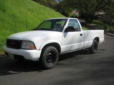 1995 Ford Bronco - This white GMC pickup truck is seen from a 3/4 front angle on a paved surface with a grassy hill behind it.