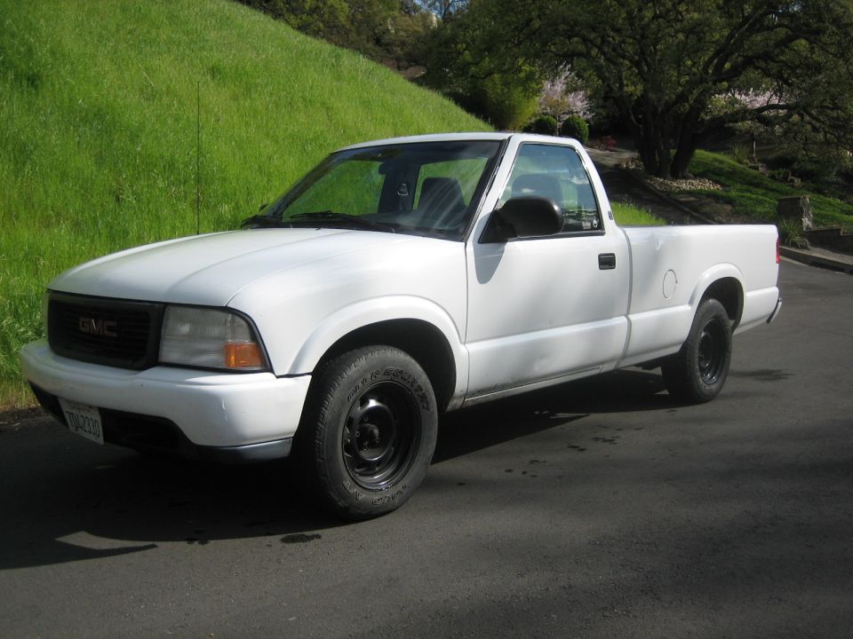 White GMC pickup truck, 3/4 front view, parked on asphalt with grassy hill in background