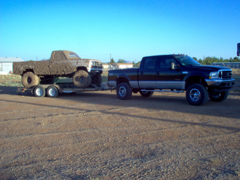 1995 Ford Bronco - A black pickup truck tows a mud-covered pickup truck on a trailer under a clear blue sky.