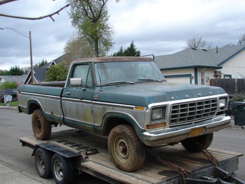 1996 Ford Bronco - A weathered, faded teal/green Ford pickup truck with heavy surface rust and patina, sitting on a flatbed trailer in a residential street se…