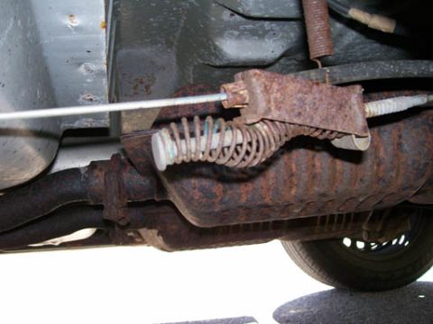 1999 Ford Crown Victoria - Close-up view of the underbody of a vehicle, showing a rusty exhaust system and a brake cable mechanism.
