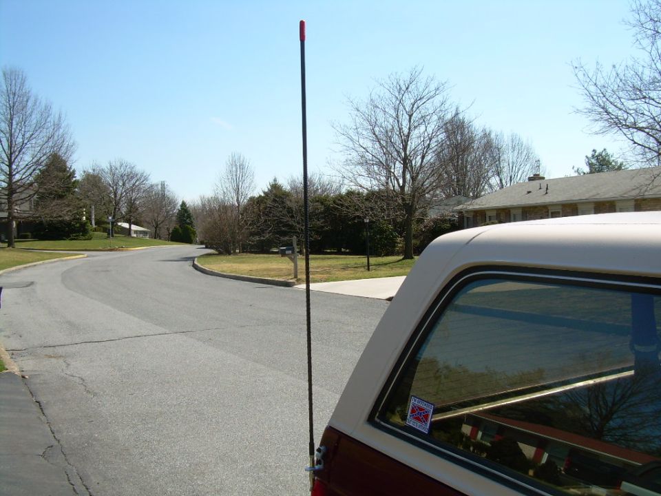 Tall black CB whip antenna mounted on the rear quarter of a red and white SUV on a suburban street