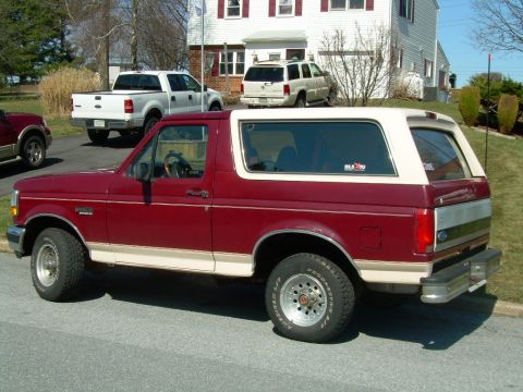 1992 Ford Bronco - A maroon and tan Ford Bronco is parked on a street with a white house and other vehicles in the background.