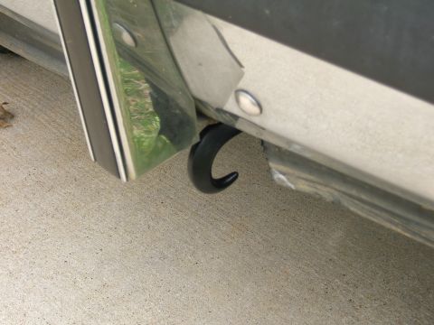 1991 Ford Bronco - Low close-up of a black J-hook tow hook poking out through a rough-cut opening in a light-colored front bumper, with a chrome trim strip an…