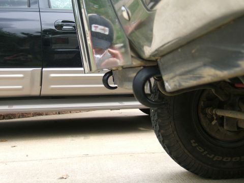 1991 Ford Bronco - Low ground-level view under the front of a truck showing a pair of black round tow hooks bolted through the frame horns behind the front bu…
