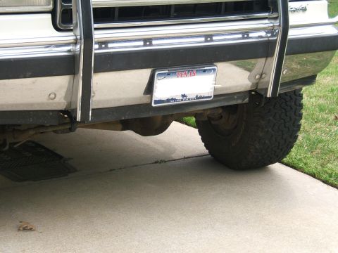1991 Ford Bronco - Low-angle view under the front of a white truck on a concrete driveway, showing the chrome front bumper, chrome tube grille guard uprights,…