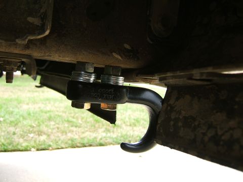 1991 Ford Bronco - Close-up from below of a black forged tow hook stamped 'MAX LOAD 10000 LB' bolted to the frame rail with two large hex bolts. The surroundi…
