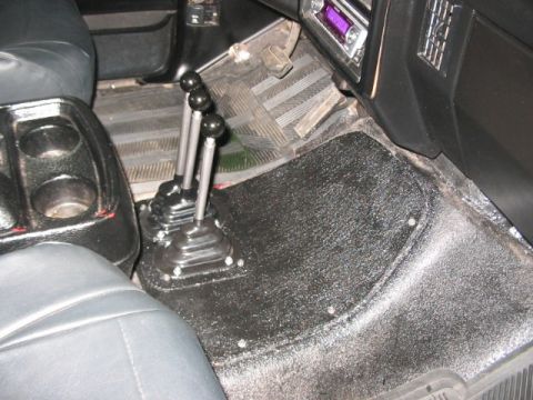 1991 Ford Bronco - The interior of a vehicle displays a center console with cup holders and three manual gear shifters, along with black textured flooring.