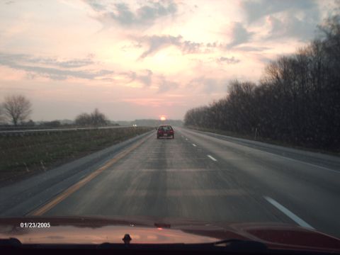 1995 Ford Probe - A lone car travels down a highway as the sun sets, casting a warm glow across the sky.