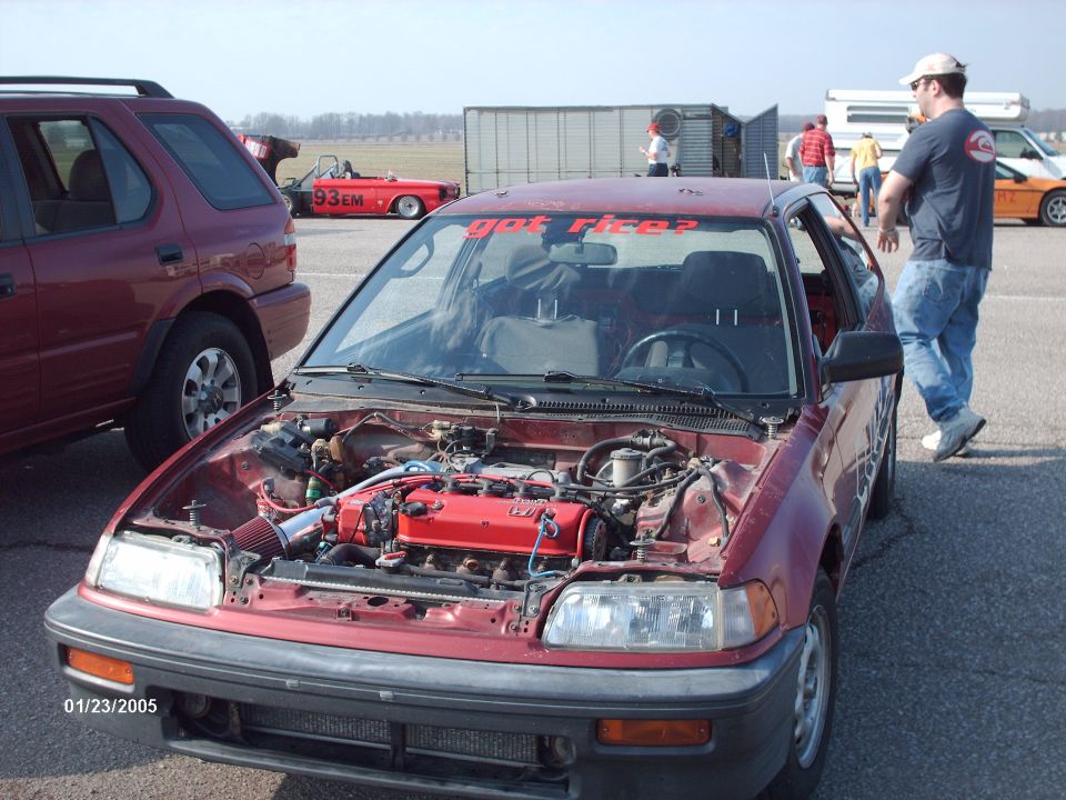 Red hatchback with hood open showing engine bay. "got rice?" sticker on windshield. Other vehicles and people in background