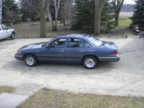 1996 Ford Crown Victoria - A blue sedan is parked on a gravel driveway next to a silver pickup truck.