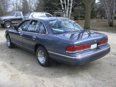 1996 Ford Crown Victoria - A blue sedan with aftermarket wheels is parked outdoors, with other vehicles in the background.