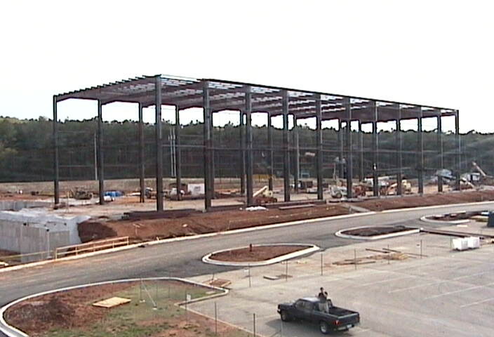 Steel frame of a large building under construction, with a pickup truck in the foreground parking lot