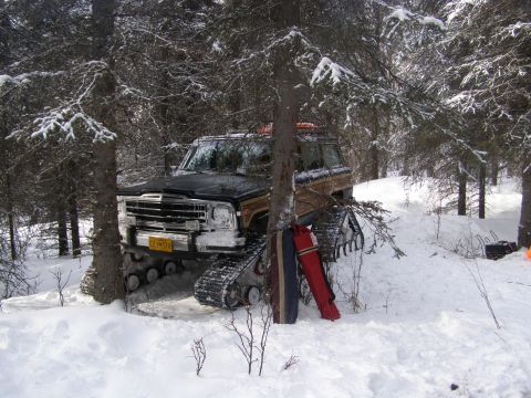 2000 Ford E350 - A Jeep Grand Wagoneer equipped with tracks is stuck in the snow, leaning against a tree in a wooded area.