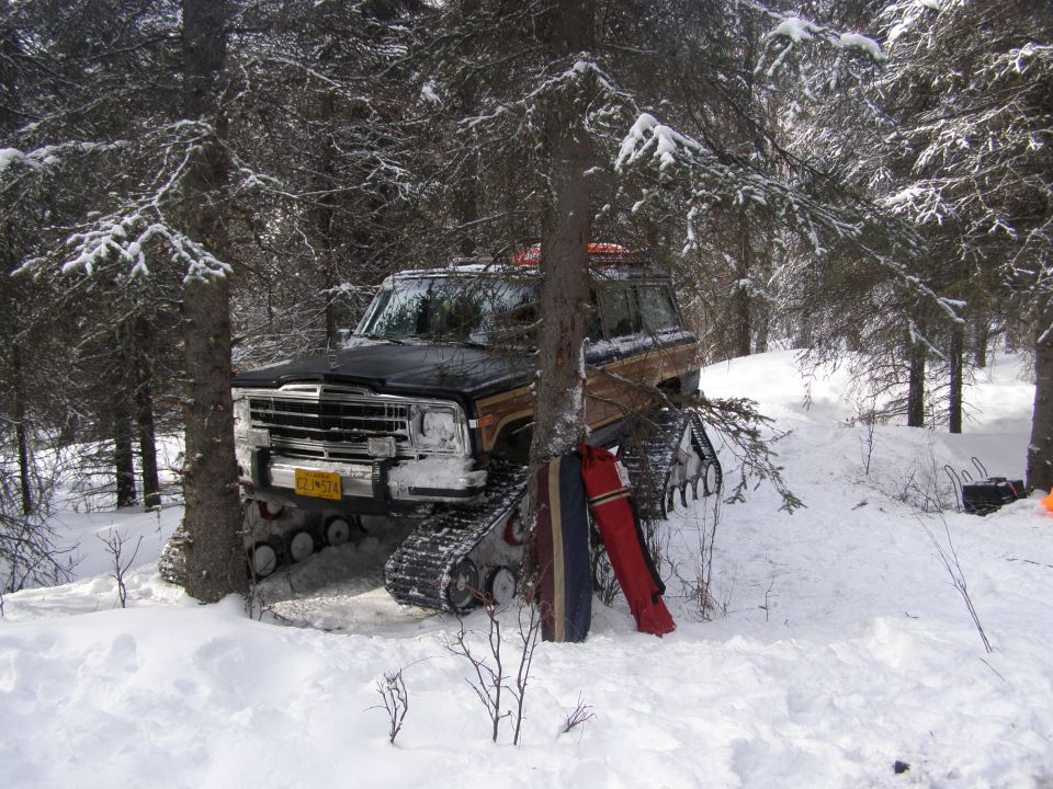 Jeep Grand Wagoneer with tracks in snow, leaning against a tree. Red and blue skis are propped up next to it