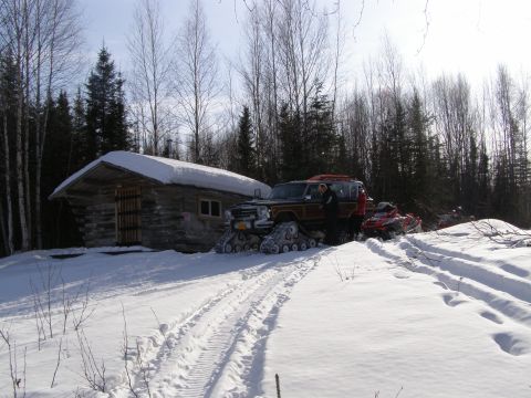 2000 Ford E350 - A Jeep Grand Wagoneer equipped with tracks sits in the snow beside a rustic cabin, with a snowmobile in the background.