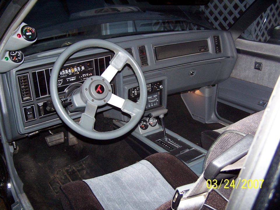 Car interior with gray dashboard, aftermarket steering wheel, and bucket seats. Gauges are mounted on the A-pillar