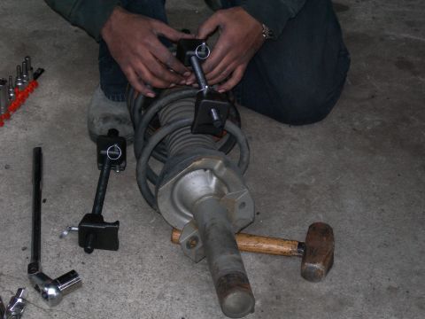 1992 Ford Taurus - A person uses a spring compressor tool to work on a coilover shock assembly, with various tools laid out on the concrete floor.