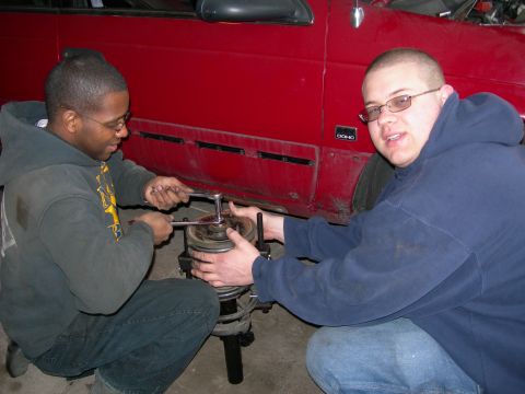 1992 Ford Taurus - Two men are shown working on a car's suspension system, with a red vehicle and its side panel visible.