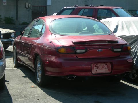 1992 Ford Taurus - This red Ford Taurus sedan is parked in a lot, partially obscured by other vehicles.