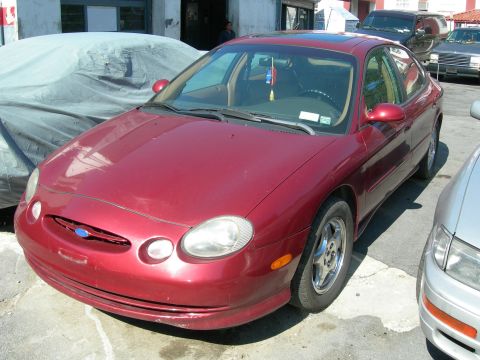 1992 Ford Taurus - This red sedan is parked on a paved surface, with other vehicles nearby and a building in the background.