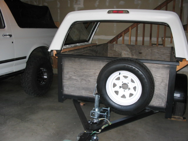 DIY trailer made from white truck cap and plywood with spare tire, beside white lifted Bronco in garage.