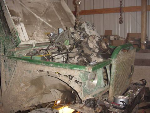 1992 Ford Bronco - A V8 engine hangs from a chain hoist over the open engine bay of a heavily mud-caked green truck inside a metal shop building. The truck's…