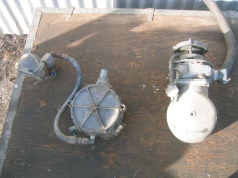 1973 Ford F-100 - Two used LPG/propane fuel-system components laid out on a weathered plywood bench, at left a round finned converter/vaporizer with a hose a…