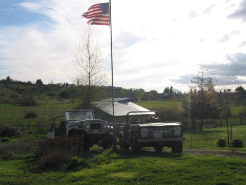 1968 Ford Bronco - Two classic off-road vehicles are parked in a grassy area next to a barn under a cloudy sky.