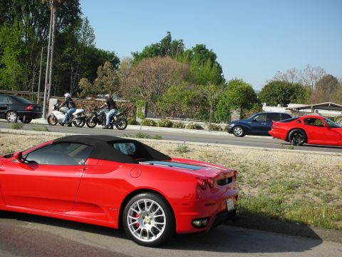 1998 Ford Crown Victoria - red Ferrari convertible parked at a roadside with its top up, while two motorcyclists and a red coupe pass on the highway behind.