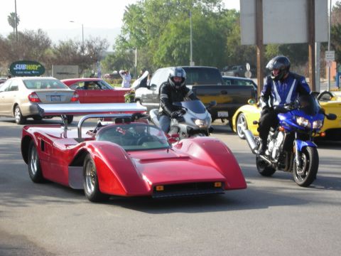 1998 Ford Crown Victoria - red open-cockpit racing-style car with a large rear wing on a public street, flanked by two motorcyclists near a Subway sign.