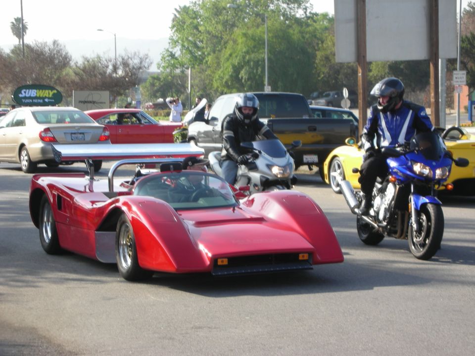 Red open-cockpit race car with rear wing on a street beside motorcycles and a Subway sign