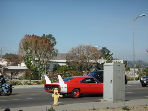 1998 Ford Crown Victoria - red winged 'Daytona' muscle car with its tall rear wing driving along a road, passing a yellow fire hydrant and a motorcyclist.