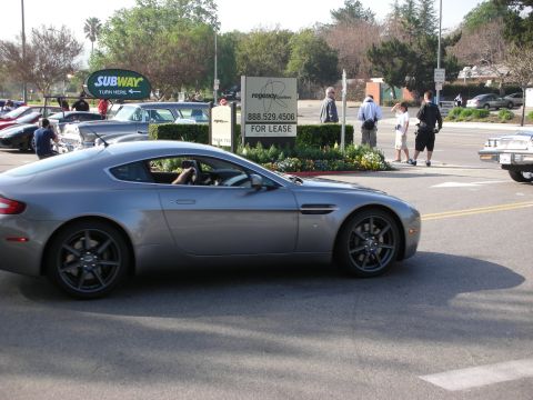1998 Ford Crown Victoria - silver-gray Aston Martin coupe driving through a street intersection, with a Subway sign and pedestrians in the background.