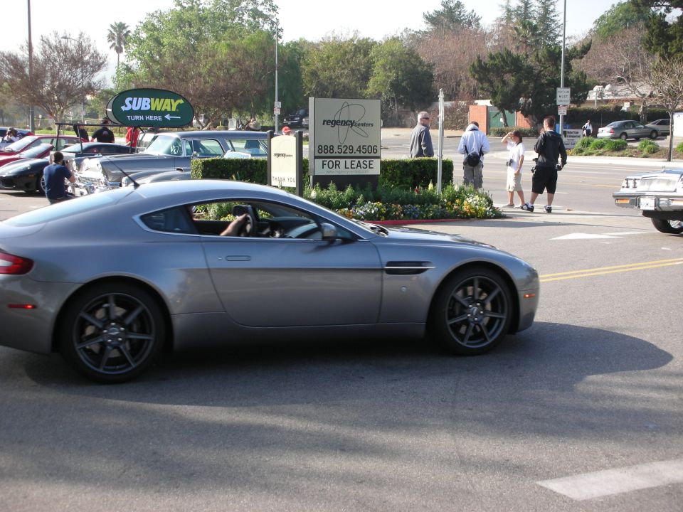 Silver Aston Martin coupe driving through an intersection near a Subway sign