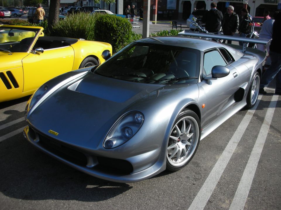 Silver Noble mid-engine sports car parked beside a yellow car at a meet