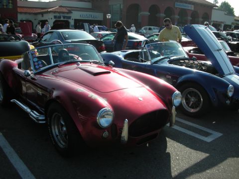 1998 Ford Crown Victoria - maroon Shelby Cobra-style open roadster sits in a sunny parking-lot car show next to a blue Cobra and a crowd of spectators.