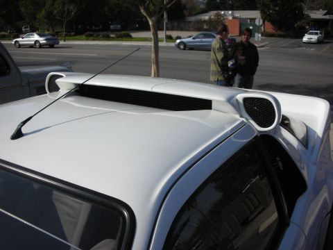 1998 Ford Crown Victoria - Close-up of a white car's roofline and rear deck showing a raised spoiler and louvered intake vents, with a street scene behind.