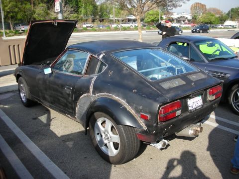 1998 Ford Crown Victoria - weathered matte-black Datsun Z coupe sits with its hood up at a parking-lot car meet, showing patchy primer and aftermarket wheels.