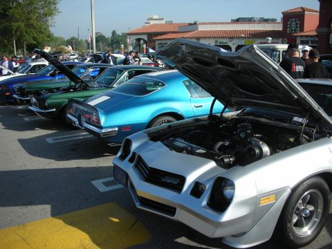 1998 Ford Crown Victoria - row of classic muscle cars with hoods raised at a car show, led by a silver Camaro and including a blue Pontiac Firebird.