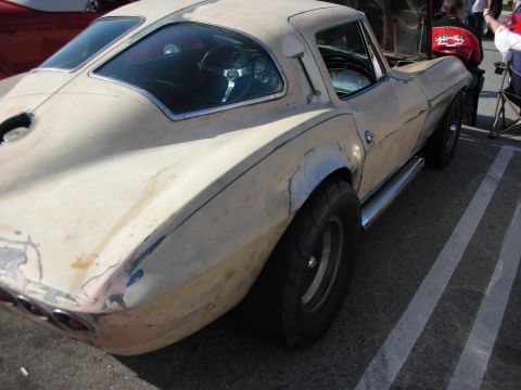 1998 Ford Crown Victoria - weathered cream fastback sports car with bare-metal and primer patches and side exhaust sits in a show parking lot.