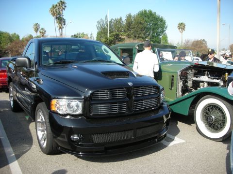 1998 Ford Crown Victoria - black Dodge Ram pickup with a hood scoop and crosshair grille parked next to a green vintage open-top car at a show.