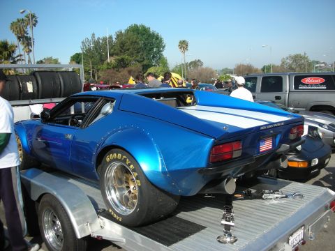 1998 Ford Crown Victoria - blue De Tomaso Pantera with white stripe and gold racing wheels sits strapped onto an aluminum trailer at a car meet.