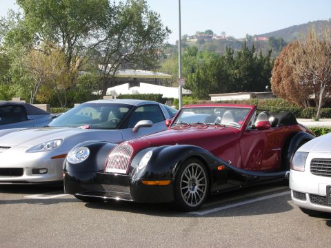 1998 Ford Crown Victoria - maroon-over-black Morgan Aero convertible sits between a silver Corvette and other cars in a sunny parking lot.